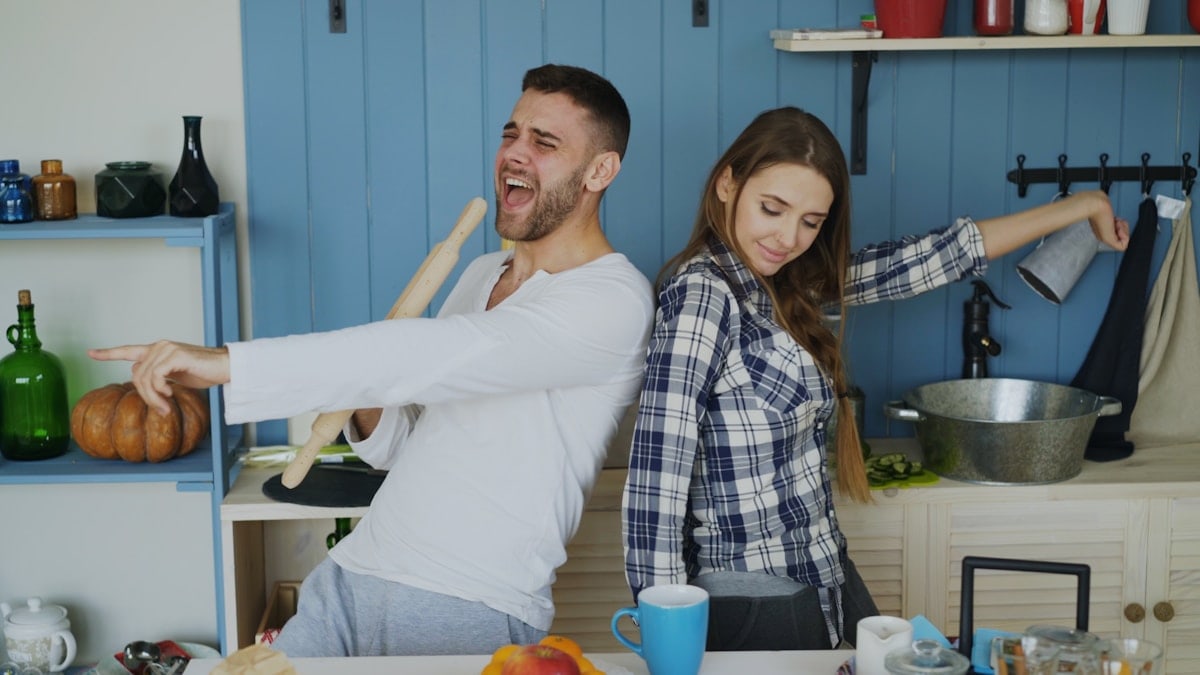 Attractive young joyful couple have fun dancing and singing while set the table for breakfast in the kitchen at home